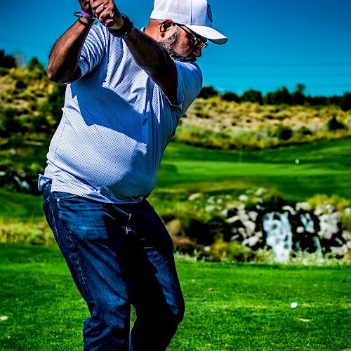 A person wearing a white cap and shirt, swinging a golf club on a lush green golf course under a clear blue sky.