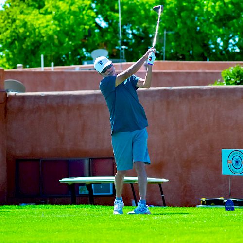 A person wearing a cap, shirt, and shorts is swinging a golf club on a grassy area with a stucco wall and green trees in the background.