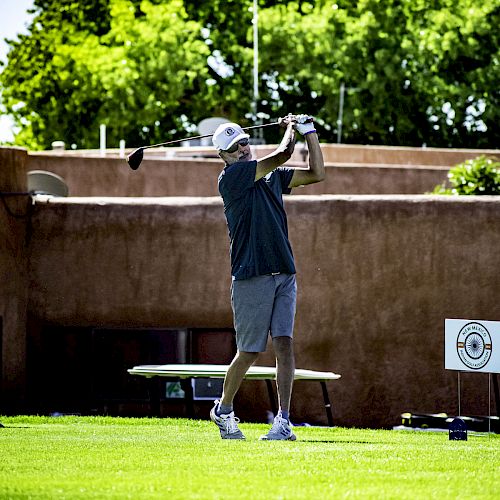 A person is playing golf, mid-swing on a green course with a backdrop of trees and a building. The player wears a cap, shorts, and a polo shirt.