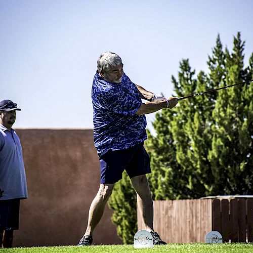 A person is swinging a golf club on a green, while another person in a hat stands nearby watching. Trees and a building are in the background.