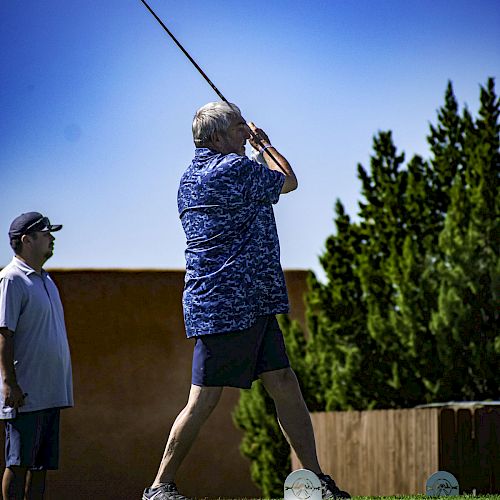 A person is swinging a golf club on a green course while another person watches. Trees and buildings are visible in the background.