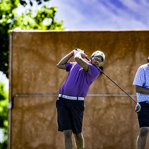 A person in a purple shirt and black shorts is swinging a golf club on a sunny day, with another person standing nearby.