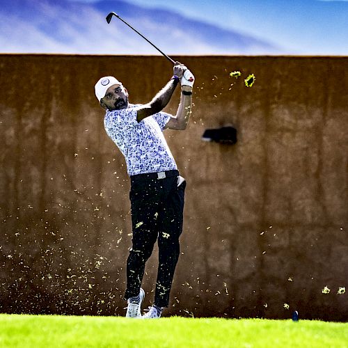 A golfer is mid-swing, with grass flying, against a backdrop of mountains. He wears a cap, patterned shirt, and dark pants.