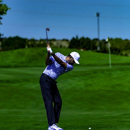 A person is playing golf, captured mid-swing on a lush green golf course under a clear blue sky, with a flag in the background.