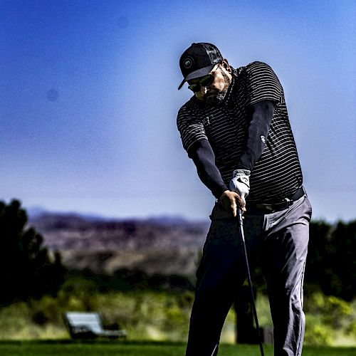 A person is swinging a golf club on a lush green golf course with mountains and a clear blue sky in the background.