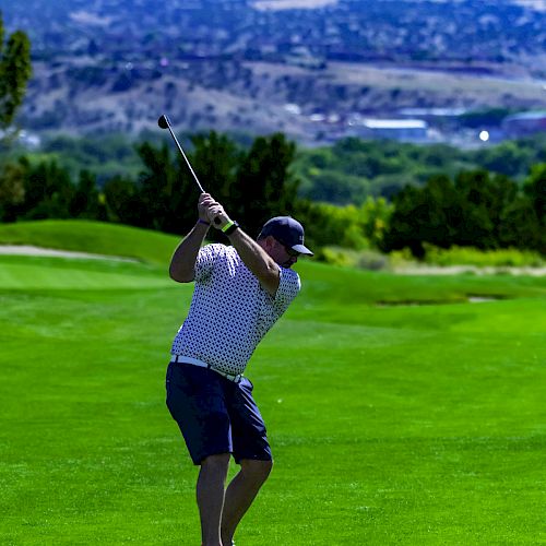 A person is playing golf on a lush green course with a picturesque mountain and valley in the background, holding a golf club mid-swing.