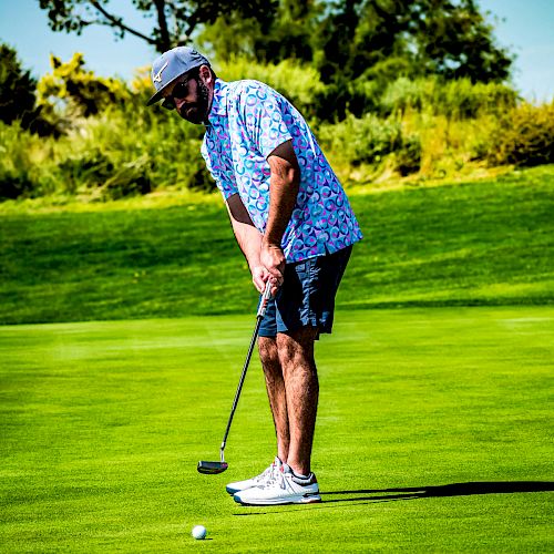 A man in casual golf attire is putting on a green golf course, with trees and shrubbery in the background.