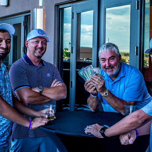 Four men are smiling and posing around a table, one holding a stack of cash. They are wearing casual attire and baseball caps.