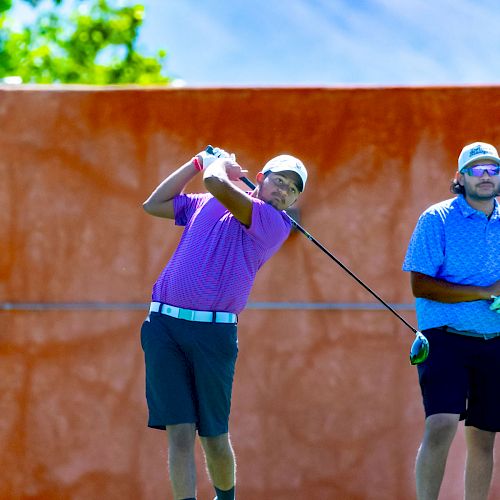 Two individuals on a golf course, one is swinging a club while the other watches. They are both dressed in golf attire.