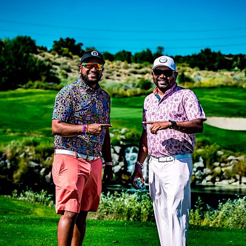 Two men in golf attire pose on a golf course with a scenic background, pointing their index fingers outward.