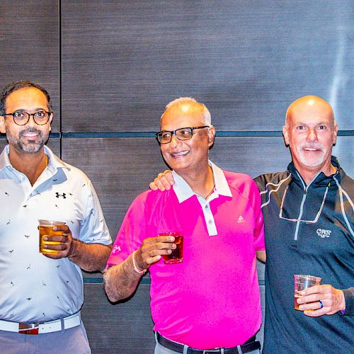 Three men are standing together, smiling, and holding drinks in a casual setting. They appear to be enjoying a social event.