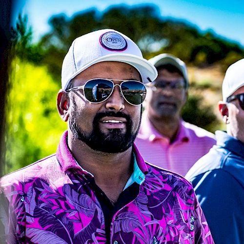 Three men are outdoors, wearing caps and sunglasses, standing in a sunny, green environment with trees and bushes in the background.