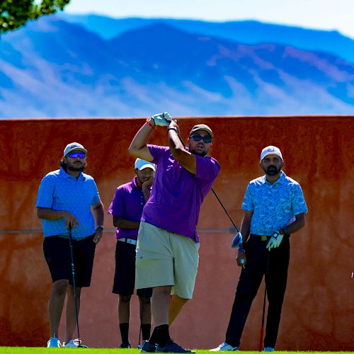 A group of four people are golfing on a sunny day, with mountains and trees in the background. One person is swinging a golf club.