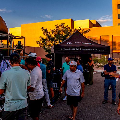 People are gathered near a food truck and tent, engaging in various activities, outside a building during sunset.