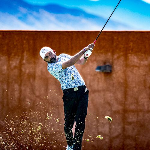 A golfer swings his club, striking the ball mid-air with a mountain backdrop, grass flying in the foreground, on a sunny day.