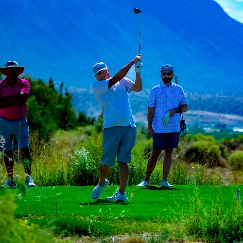 Three people are on a golf course with mountainous scenery in the background. One person is swinging a golf club while the other two watch.