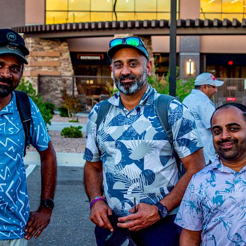 Three men wearing casual shirts, standing outside a building, smiling, and posing for a photograph.