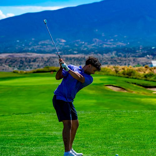 A person is playing golf on a scenic course with a mountain in the background, preparing to hit the ball with a club on a sunny day.