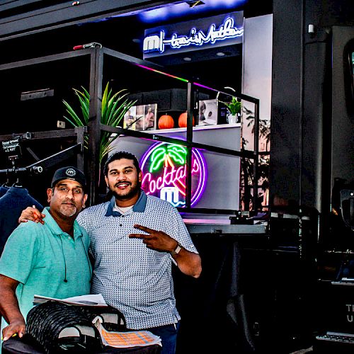 Two people are posing in front of a mobile retail truck displaying clothing. A neon 