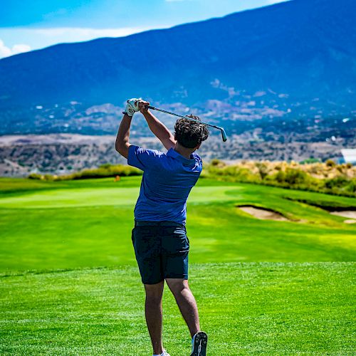 A person is playing golf on a lush green course, swinging a club with mountains and blue sky in the background.