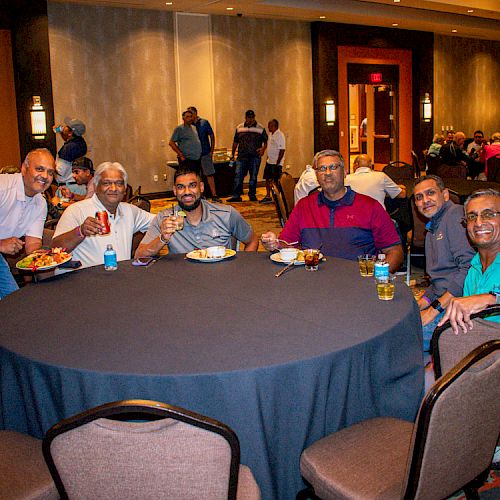 A group of six people are sitting around a table in a banquet hall, smiling and holding drinks. Other people are visible in the background.