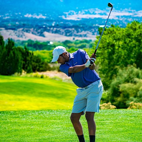 A person is swinging a golf club on a lush golf course with a scenic mountain backdrop, wearing a blue shirt, white shorts, socks, and a white cap.