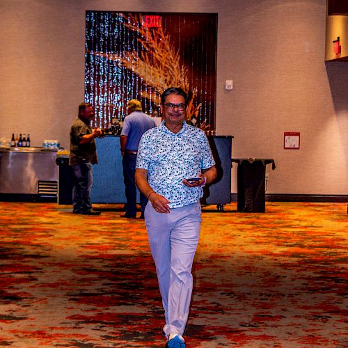 A man is walking in a spacious, well-lit room with a colorful carpet and modern decor, holding a phone. People are gathered near a table in the background.