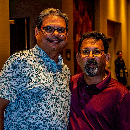 Two men standing together in a dimly lit hallway or room, one smiling and the other with a neutral expression.