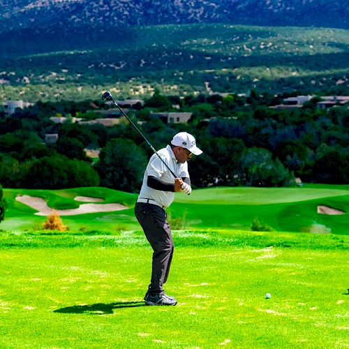 A golfer in mid-swing on a lush green course, with rolling hills and mountains in the background, surrounded by trees.