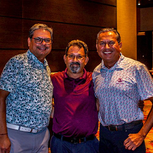 Three men are standing indoors, posing for a photo and smiling at the camera. They are dressed in casual attire.