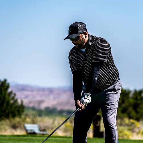 A person is getting ready to take a golf swing on a golf course, with a scenic background of trees and hills visible under a clear sky.