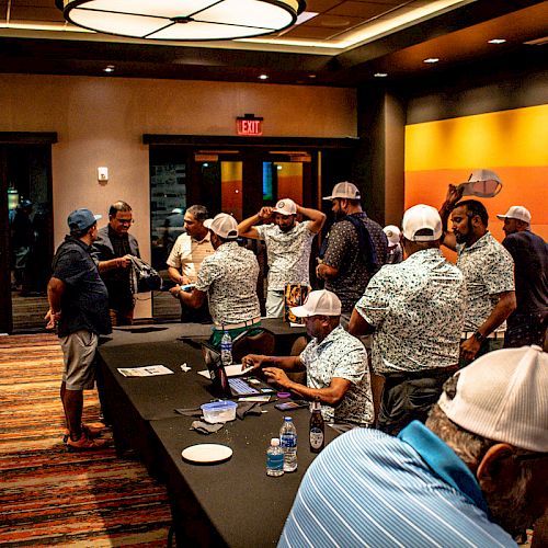 Several people are gathered in a room, some wearing matching shirts and hats, likely attending an event or meeting with tables and a presentation screen.