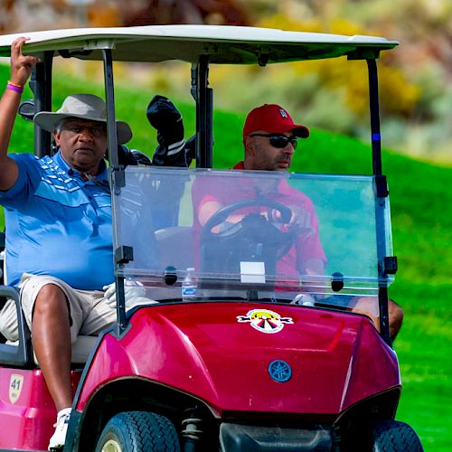 Two people are riding in a red golf cart on a golf course. The person on the left is waving while the person on the right is driving the cart.