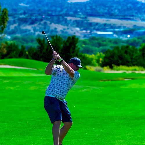 A person is swinging a golf club on a lush green golf course with a scenic background of hills and a clear blue sky visible in the distance.