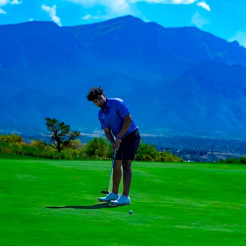 A person is playing golf on a well-maintained green, with a mountainous backdrop under a bright, partly cloudy sky.