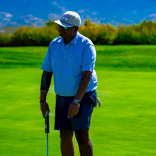 A person is standing on a golf course, holding a golf club, with mountains and greenery in the background.