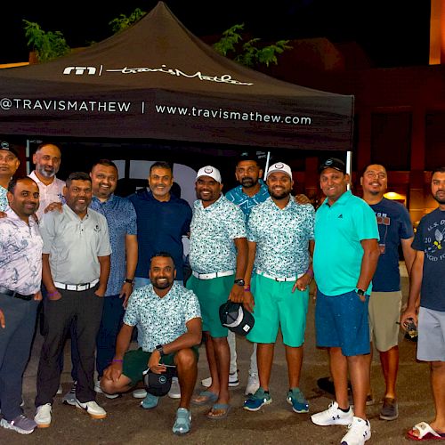 A group of people pose under a TravisMathew tent at night, dressed casually, with some in matching outfits, outside a building with illuminated lights.