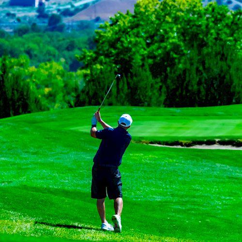 A person is playing golf on a lush green course, mid-swing, with trees and hills in the background.