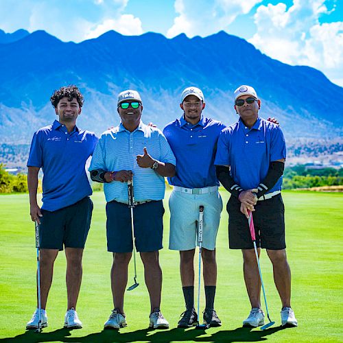 Four people standing on a golf course with mountains in the background, each holding golf clubs and smiling at the camera.