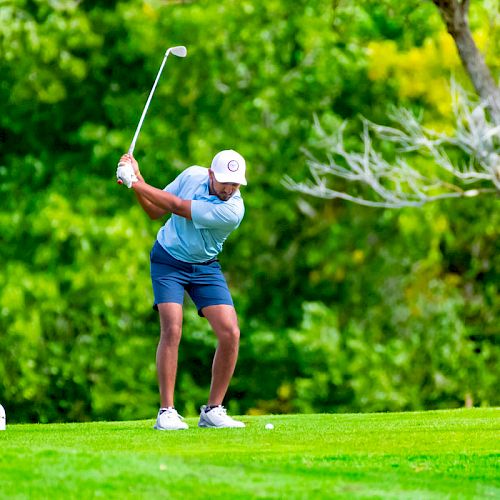 Two men are on a golf course. One man is swinging a golf club while the other stands nearby. They are surrounded by lush green trees.