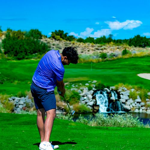 A person wearing a blue shirt and shorts is playing golf on a lush green course with a small waterfall in the background.