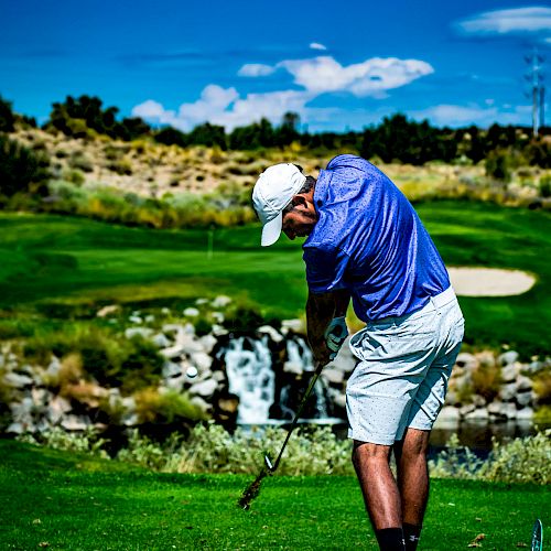 A person in a blue shirt and white shorts, swinging a golf club on a green golf course with a waterfall in the background, under a clear blue sky.