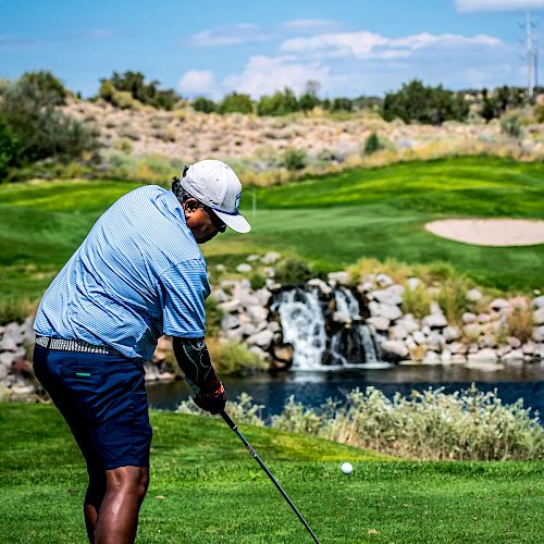 A person in a golf outfit is mid-swing on a golf course with a waterfall and green landscape in the background.