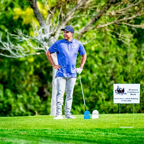 A person in a blue shirt and gray pants standing on a golf course, holding a golf club, with a sign nearby, surrounded by greenery.