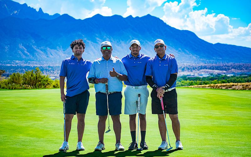 Four men on a golf course posing with mountains in the background, wearing blue shirts and holding golf clubs, on a sunny day, smiling for the camera.
