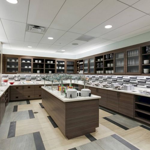 A modern kitchen or buffet area with shelves full of plates, cups, and utensils, featuring a central island and tiled walls.