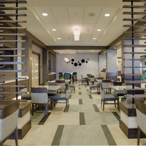 The image shows a modern dining area with tables, chairs, and decorative partitions, featuring a neutral color palette and stylish lighting.