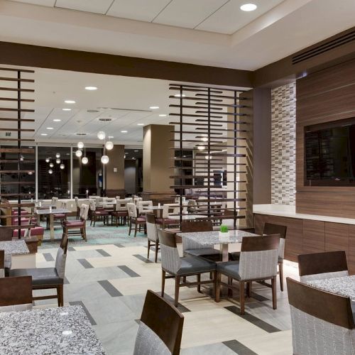 A modern restaurant interior with dining tables, chairs, decorative panels, and a wall-mounted TV. Soft lighting creates a cozy atmosphere.