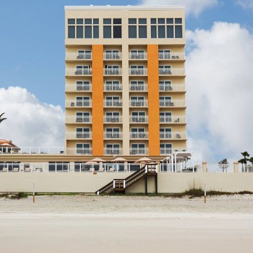 A beachfront hotel with multiple floors and balconies, featuring palm trees and a sandy beach in the foreground.