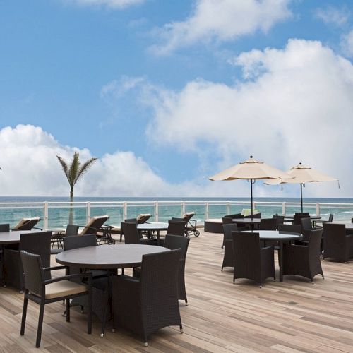 Outdoor patio with tables, chairs, and umbrellas overlooking the ocean. Few palm trees are visible under a blue sky with white clouds.